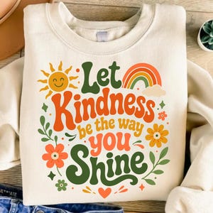 May include: A cream-colored crewneck sweatshirt featuring a graphic print with the text Let Kindness be the way you SHINE. The design includes a yellow sun, a multi-colored rainbow, orange and yellow flowers, green leaves, and a small orange heart.