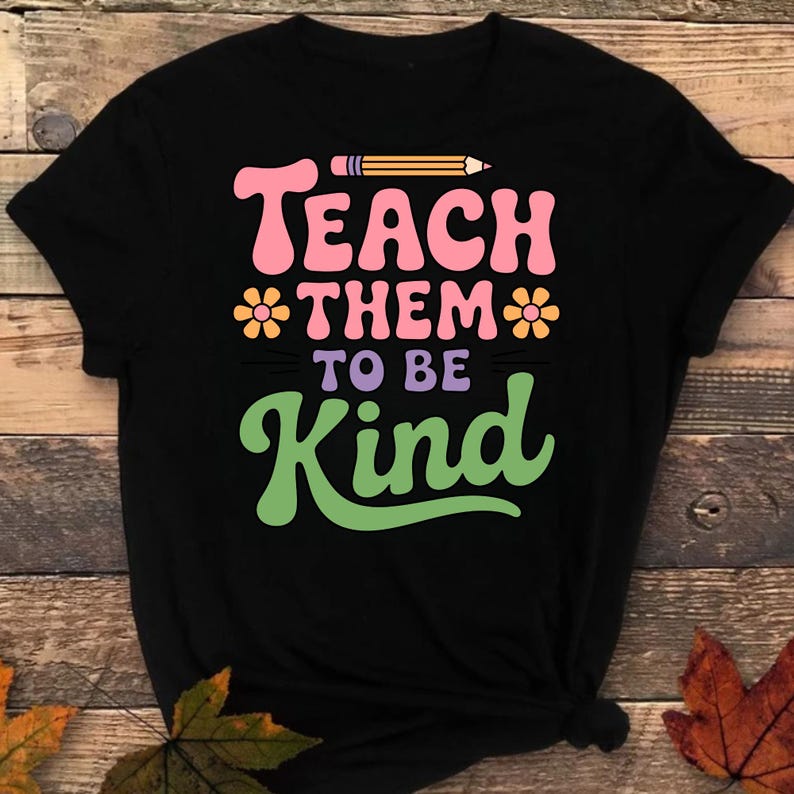 May include: Black t-shirt with the words "Teach Them To Be Kind" in pink, purple, and green, with a pencil graphic. The shirt has a crew neck and short sleeves.