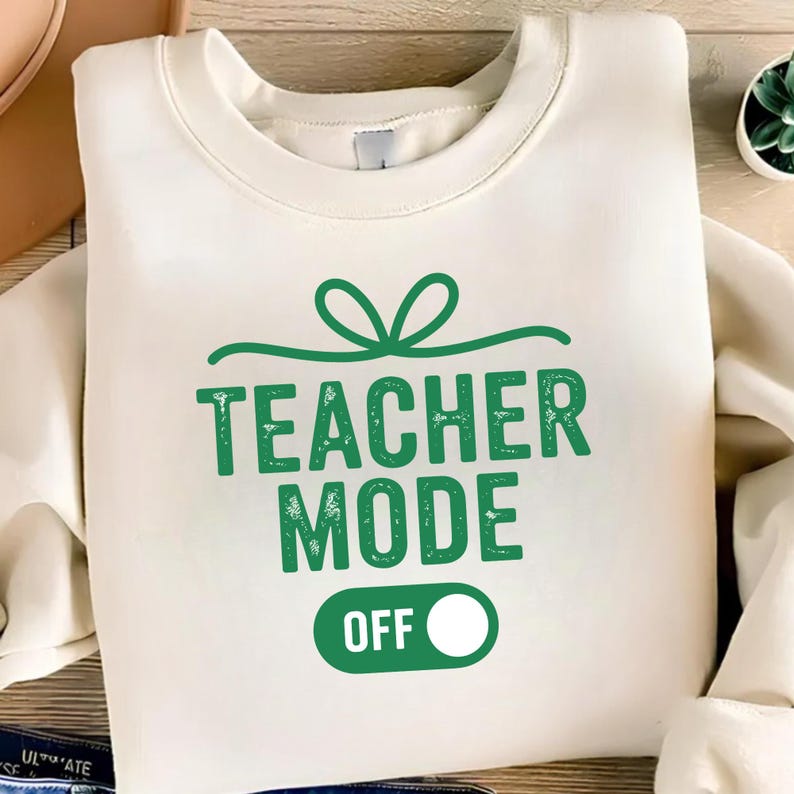 May include: Cream-colored sweatshirt with the text "TEACHER MODE OFF" in green. The design includes a green bow above the text and a green switch graphic with a white circle indicating "OFF".