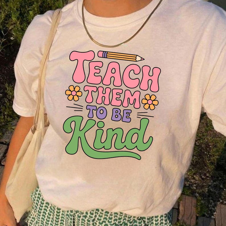 May include: White t-shirt with the text "Teach Them To Be Kind" in pink, purple, and green. The design includes a pencil and flower graphics. The shirt has a crew neck and short sleeves.