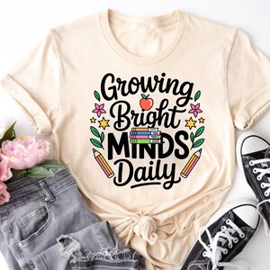 May include: A cream-colored t-shirt with the text "Growing Bright Minds Daily" in black script. The design includes a red apple, books, flowers, stars, and pencils. The shirt is paired with gray jeans and black canvas shoes.