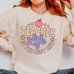 May include: Cream-colored sweatshirt with the text "TEACHER Life IS THE BEST LIFE" in a retro font. The design includes an apple, stars, and flowers in shades of pink, peach, and purple. A cute and stylish design for teachers.