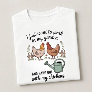 May include: White t-shirt featuring a whimsical illustration of two chickens, a fence, and a watering can. The text reads, "I just want to work in my garden AND HANG OUT with my chickens."