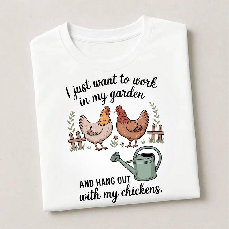 May include: White t-shirt featuring a whimsical illustration of two chickens, a fence, and a watering can. The text reads, "I just want to work in my garden AND HANG OUT with my chickens."