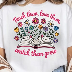 May include: White t-shirt with a colorful floral design and the text "Teach them, love them, watch them grow." The graphic features an open book with flowers in various colors, including pink, yellow, blue, and purple.