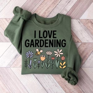 May include: An olive green sweatshirt with the text "I LOVE GARDENING" in black. Below the text is a colorful floral design with a bee. The flowers are in shades of blue, pink, yellow, and white.