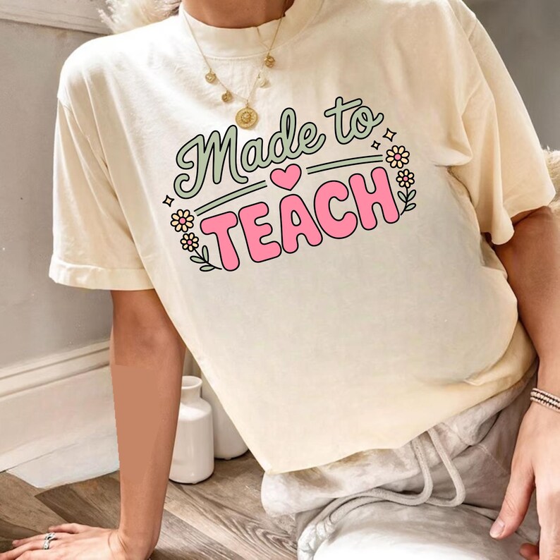 May include: Cream-colored t-shirt with the text "Made to TEACH" in a retro font. The text is in green and pink, with floral and heart accents. The shirt is a casual style.