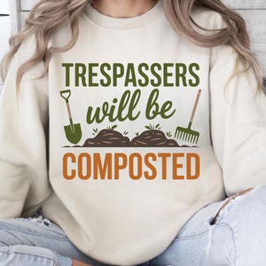 May include: Cream-colored sweatshirt with the text "Trespassers will be composted" in green and orange. The design includes a shovel, soil mounds, and a rake, all in green, evoking a gardening theme.