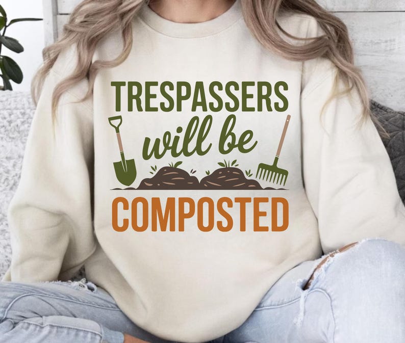 May include: Cream-colored sweatshirt with the text "Trespassers will be composted" in green and orange. The design includes a shovel, soil mounds, and a rake, all in green, evoking a gardening theme.