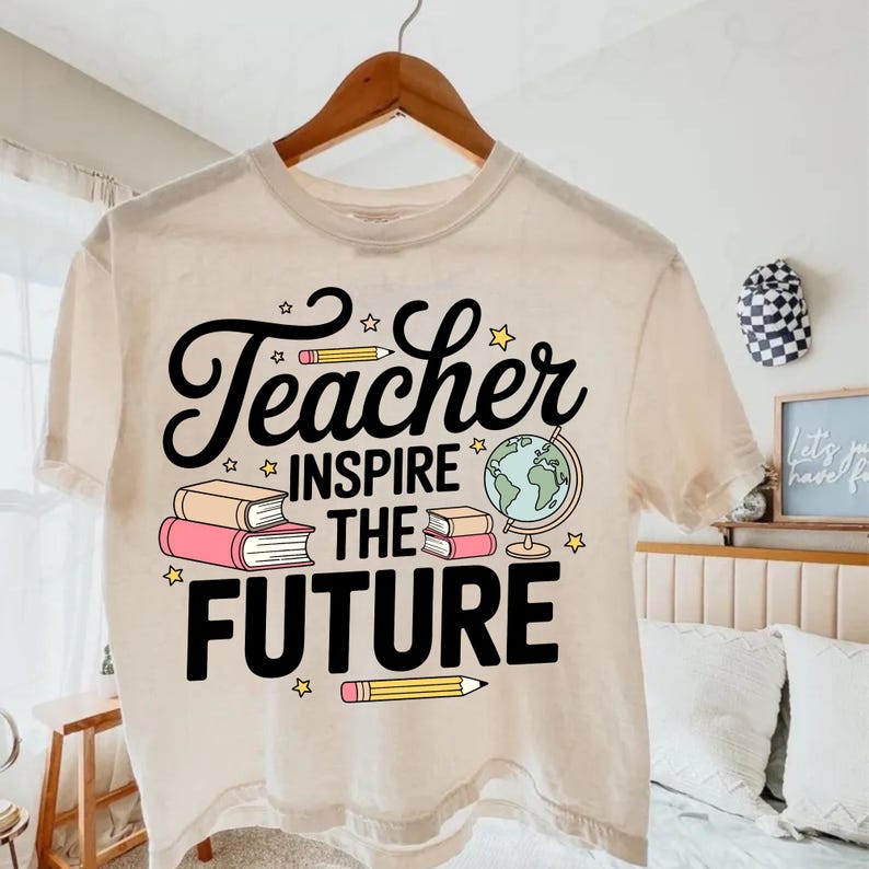 May include: A beige t-shirt with the words "Teacher Inspire the Future" in black script and block letters. The design includes illustrations of books, pencils, stars, and a globe. The shirt is hanging on a wooden hanger.
