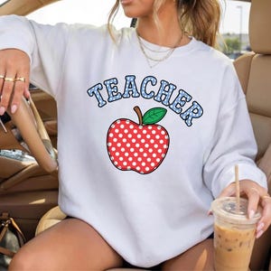 May include: White sweatshirt featuring the word "TEACHER" in blue polka dot letters above a red apple with white polka dots. The apple has a green leaf and a brown stem. The sweatshirt is a casual style.