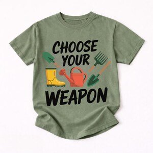 May include: Olive green t-shirt with the text "CHOOSE YOUR WEAPON" in black. The design includes illustrations of gardening tools: a shovel, a fork, a watering can, and a yellow rain boot.