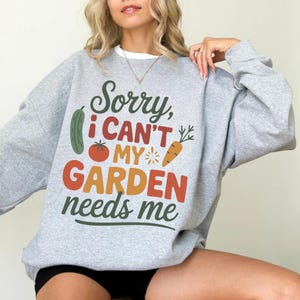 May include: A light gray sweatshirt with the text "Sorry, I can't, my garden needs me" in green, red, and orange. The design includes illustrations of a cucumber, tomato, and carrot.