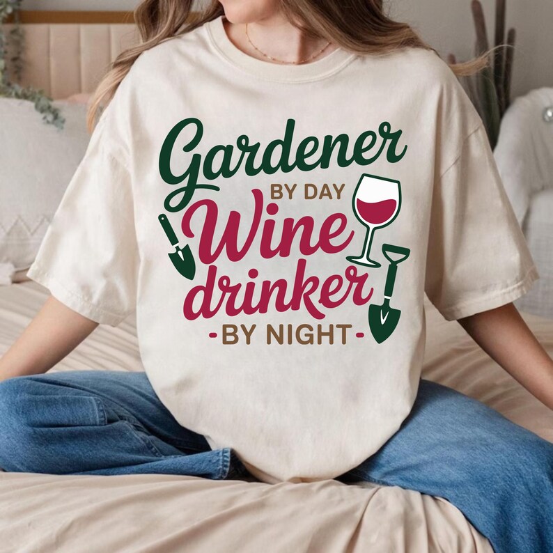 May include: Cream-colored t-shirt with the text "Gardener by Day Wine Drinker by Night" in green and burgundy. The design includes a wine glass and gardening tools. The shirt is a casual style.