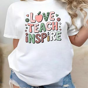 May include: White t-shirt with the words "LOVE TEACH INSPIRE" in colorful, patterned letters. The design includes an apple, flowers, and a pencil, perfect for teachers or anyone in education.