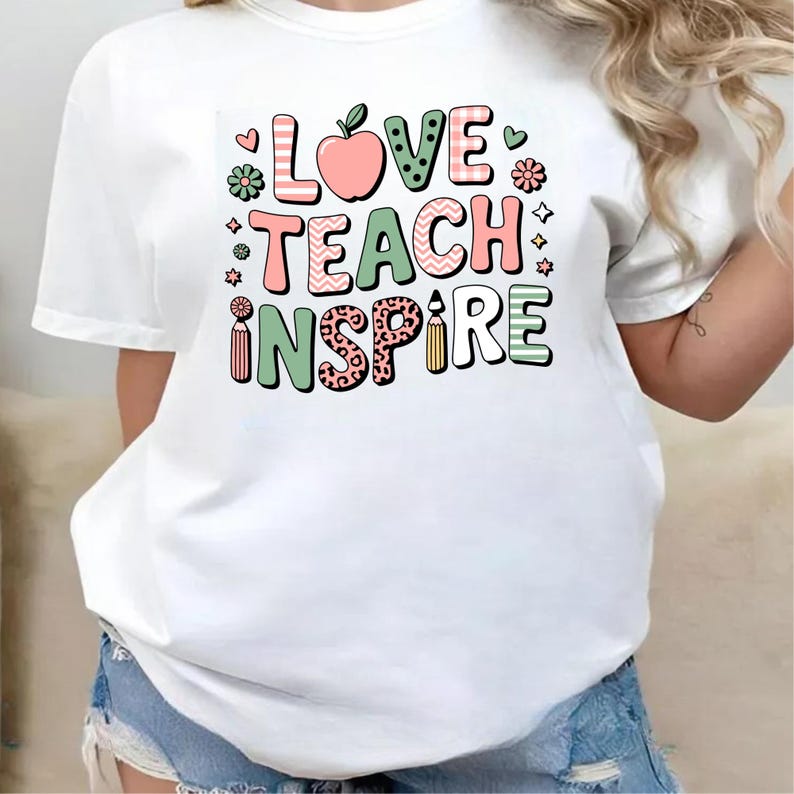 May include: White t-shirt with the words "LOVE TEACH INSPIRE" in colorful, patterned letters. The design includes an apple, flowers, and a pencil, perfect for teachers or anyone in education.