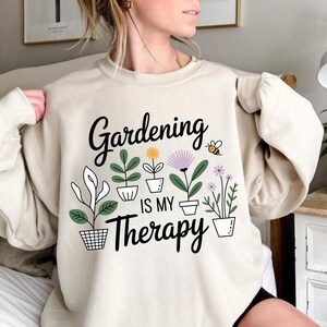 May include: Beige sweatshirt with the text "Gardening is my Therapy" in black script. The design includes potted plants and a small bee illustration. A comfortable, casual top for gardening enthusiasts.