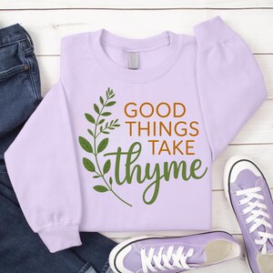 May include: A light purple sweatshirt with the phrase "GOOD THINGS TAKE thyme" in brown and green lettering. A green leafy sprig design is on the left side of the text. The sweatshirt is paired with jeans and purple sneakers.