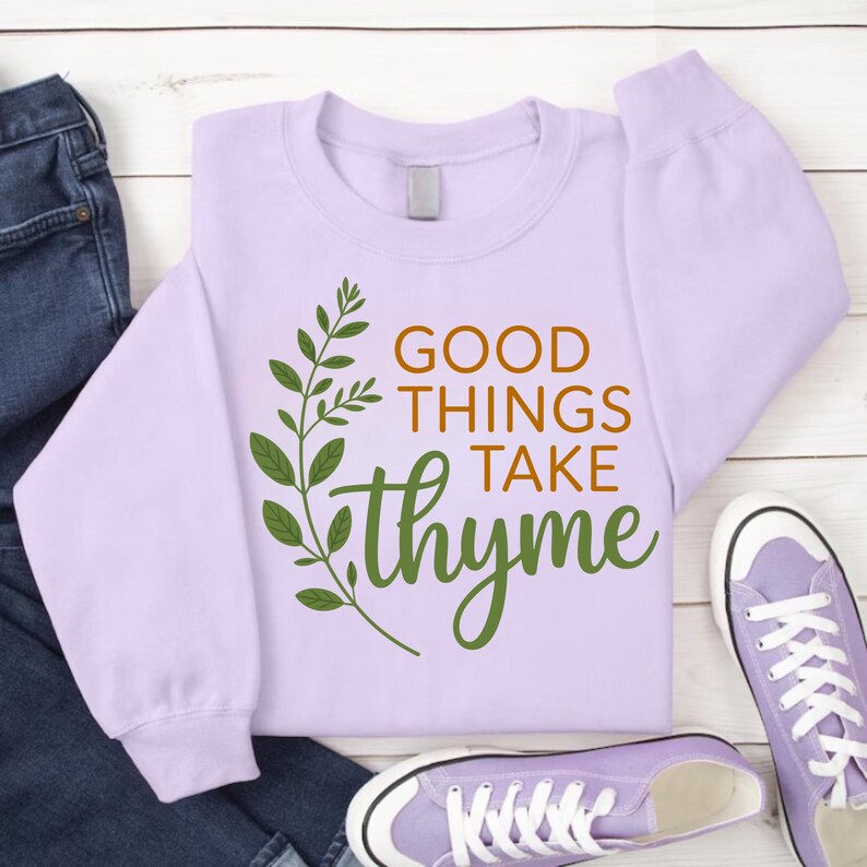 May include: A light purple sweatshirt with the phrase "GOOD THINGS TAKE thyme" in brown and green lettering. A green leafy sprig design is on the left side of the text. The sweatshirt is paired with jeans and purple sneakers.