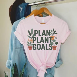 May include: A light pink t-shirt with the words "PLAN PLANT GOALS" in green and black lettering. The design includes illustrations of potted plants. The shirt is hanging on a wooden hanger with other clothing items in the background.