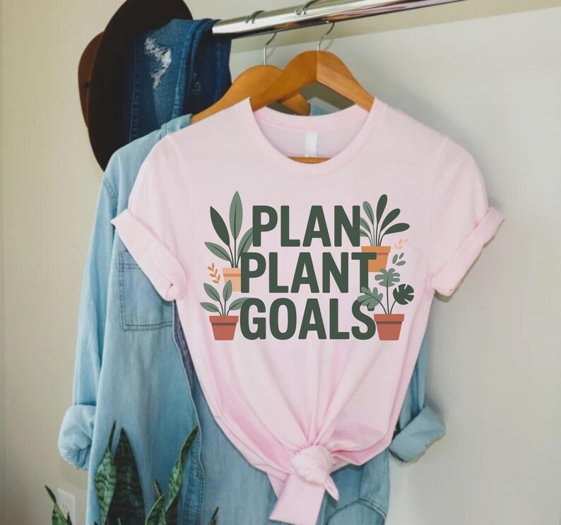 May include: A light pink t-shirt with the words "PLAN PLANT GOALS" in green and black lettering. The design includes illustrations of potted plants. The shirt is hanging on a wooden hanger with other clothing items in the background.