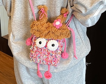 May include: A small, rectangular crochet crossbody bag featuring a textured, multicolored pink and blue yarn body. It has two large white eyes with black pupils, dangling pink yarn legs with round feet, and a brown fuzzy top with a pink flower.