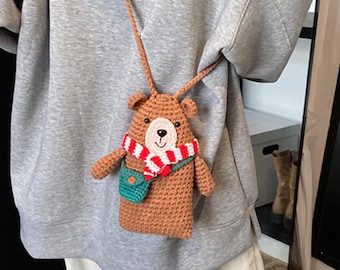 May include: A brown crocheted crossbody bag shaped like a bear. The bear features a red and white striped scarf, a small green pouch across its chest, and a smiling face with black eyes and a dark nose. It hangs from a long, thin brown strap.