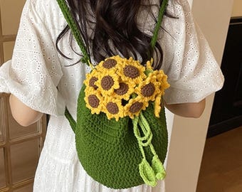 May include: A green crochet backpack featuring a cluster of yellow and brown sunflowers attached to the top. The bag has two green crochet shoulder straps and a drawstring closure with two small green leaf-shaped accents hanging from the front.