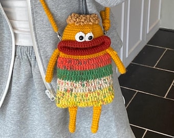 May include: A whimsical, handmade crochet crossbody bag in the shape of a cartoon character. The bag features a yellow body, orange arms and legs, and a red mouth. The bag has a striped pattern in shades of green, orange, and yellow. The strap is yellow.