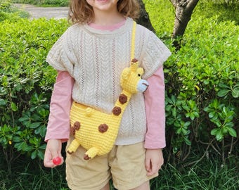 May include: A yellow crochet giraffe-shaped crossbody bag with brown spots, a long neck, and a light blue snout. The bag features a long strap and is worn over a cream-colored knit top, a pink long-sleeve shirt, and tan shorts in an outdoor setting.