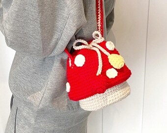 May include: A red crochet mushroom-shaped bag with white polka dots and a cream-colored base. The bag has a long red and cream strap and a cream-colored bow. The bag is a unique accessory.