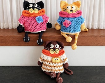 May include: Three crocheted cat dolls with googly eyes and yarn sweaters. The black cat wears a pink sweater, the orange cat wears a blue sweater, and the brown cat wears a white and orange patterned sweater. Text reads: THREE CHEEKY CATS.
