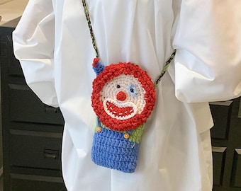 May include: A handmade crochet crossbody bag featuring a clown face design with a red yarn hair border, a white face, a red nose, and a wide smiling mouth. The bag has a blue crocheted body with green accents and a braided green and black strap.