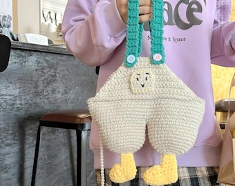 May include: A whimsical, handmade crochet bag in the shape of overalls. The bag is primarily cream-colored with teal straps and yellow feet. The bag features a small face design and is held by a person.