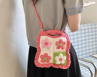 May include: A pink crocheted handbag with a floral design. The bag features four squares with pink and white flowers, and a green and white flower. It has a pink handle and strap.