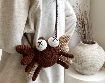 May include: A brown and tan crochet crab-shaped crossbody bag with white eyes and black cross markings. The bag has a long brown strap and tan wings. The bag is a unique accessory.