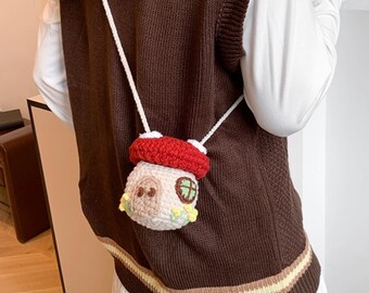 May include: A whimsical, crocheted mushroom-shaped crossbody bag. The bag features a red cap, a beige body with a small door and window, and a white strap. The bag is worn over a brown sweater.
