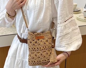 May include: A beige crochet crossbody bag featuring an openwork diamond pattern, a braided shoulder strap, and a small rectangular brown leather-like label with a crown icon. A matching beige tassel with a wooden bead hangs from the side.