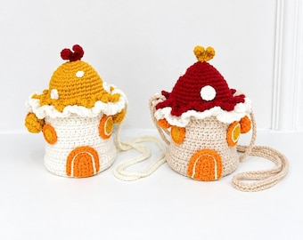 May include: Two crochet bags shaped like mushroom houses. One bag has a yellow cap with white spots and a red top detail, while the other has a red cap with white spots and a yellow top detail. Both feature cream bodies, orange doors, and long straps.