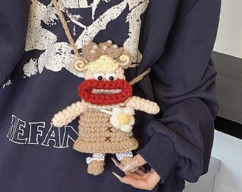 May include: A handmade crochet crossbody bag in the shape of a cartoon character. The bag features a beige body, red mouth, and a flower accent. The bag is suspended from a beige cord.