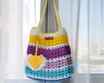 May include: A handmade crochet tote bag in a rainbow of colors, including white, yellow, purple, and blue. The bag features a heart-shaped charm and a leather tag. The bag is suspended by white and yellow straps.