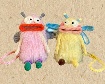 May include: Two handmade plush monster toys. One is pink with a red mouth, gray arms, and green feet. The other is yellow with a pink mouth, blue wings, and a brown foot. Both have white eyes and a loop for hanging.
