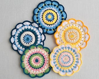 May include: Five handmade crochet coasters in various colors, including blue, yellow, green, and pink. Each coaster features a circular design with intricate patterns and scalloped edges. The coasters are arranged in a circular pattern.