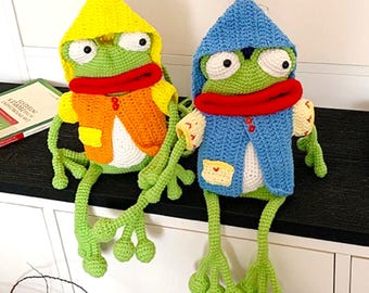May include: Two handmade crochet frog dolls with large white eyes and prominent red lips. The left frog wears a yellow hooded jacket over an orange vest. The right frog wears a blue hooded jacket over a blue vest. Both have long green limbs.