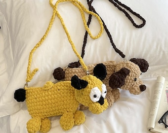 May include: Two crocheted dog-shaped crossbody bags with long braided straps. One bag is bright yellow with black ears, a black tail, and large white eyes. The second bag is beige with dark brown ears, a dark brown tail, and dark brown accents.