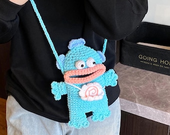 May include: A light blue crochet crossbody bag shaped like a cartoon monster with a large pink mouth and white eyes. The bag has a light blue strap and a white and pink design on the front.