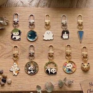 Könnte beinhalten: Eine Sammlung handgefertigter Charm-Schlüsselanhänger mit verschiedenen Designs, darunter Pandas, ein Geist und eine Katze. Die Schlüsselanhänger werden auf einem Holzbrett mit einer Karte mit der Aufschrift "Handmade Charm Collection" präsentiert.