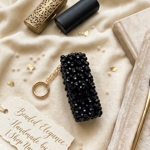 May include: A black beaded lipstick holder with a gold keychain, next to a gold ornate case and a black leather case. A tag reads "Beaded Elegance - Handmade by [Shop Name]" on a cream-coloured surface with scattered pearls.