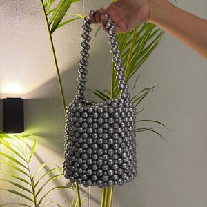May include: A gray beaded handbag with a cylindrical shape and a matching beaded handle. The bag is held up against a background of green plants and a white wall. The beads appear to be a uniform size and color.