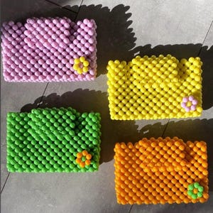 Puede incluir: Cuatro bolsos rectangulares hechos de pequeñas cuentas esféricas en rosa, amarillo, verde y naranja. Cada bolso presenta un detalle de cuenta en forma de flor en contraste en la esquina inferior derecha, creando una superficie texturizada en cuadrícula.